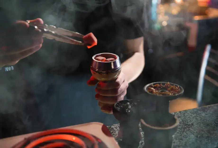 Person preparing hookah charcoals and loading shisha into a hookah bowl